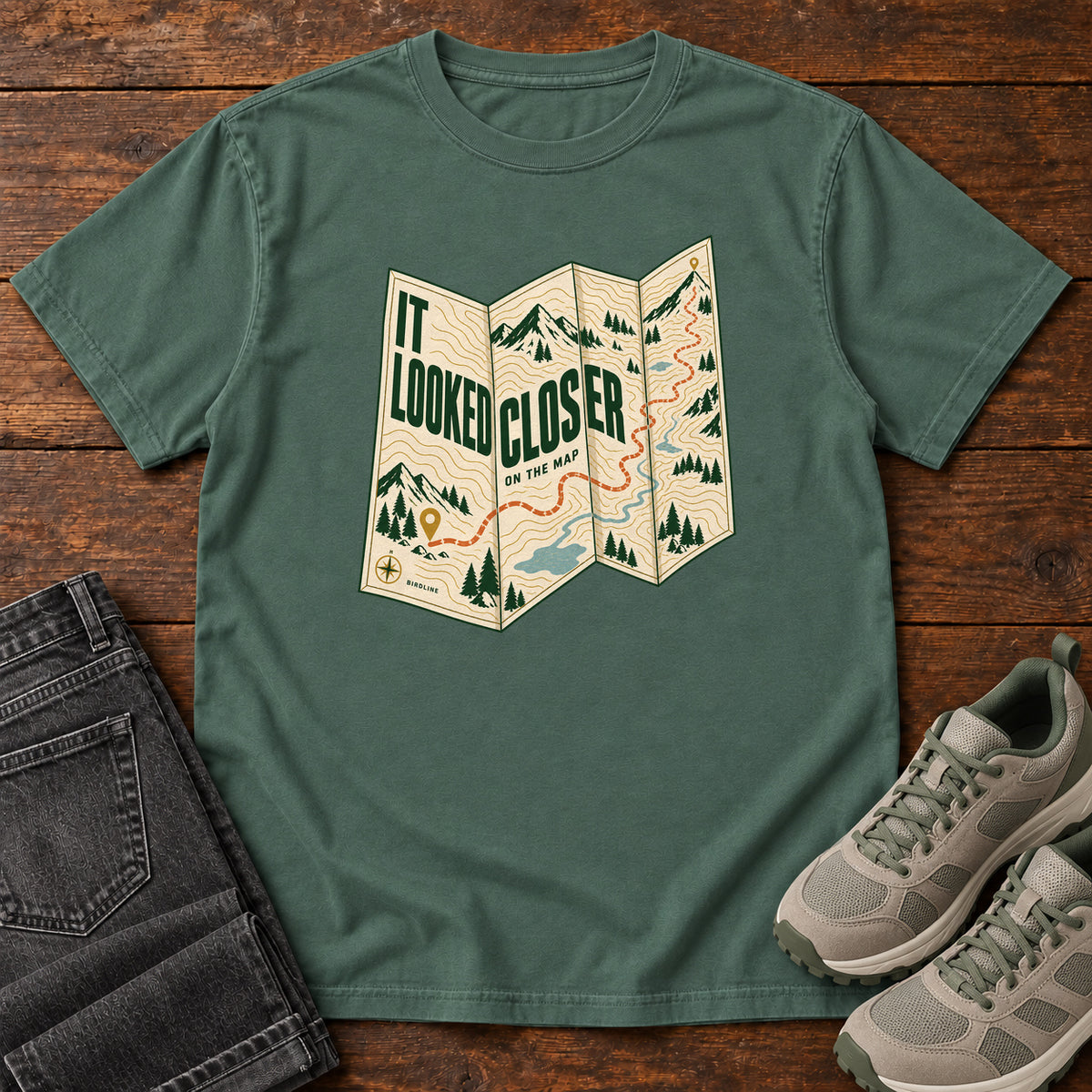 Blue Spruce It Looked Closer on the Map T-Shirt with a folded cream map graphic, green lettering and an orange route, front flat lay on wood.