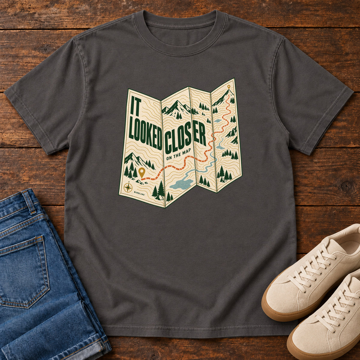 Graphite It Looked Closer on the Map T-Shirt with a folded cream map graphic, green lettering and an orange route, front flat lay on wood.