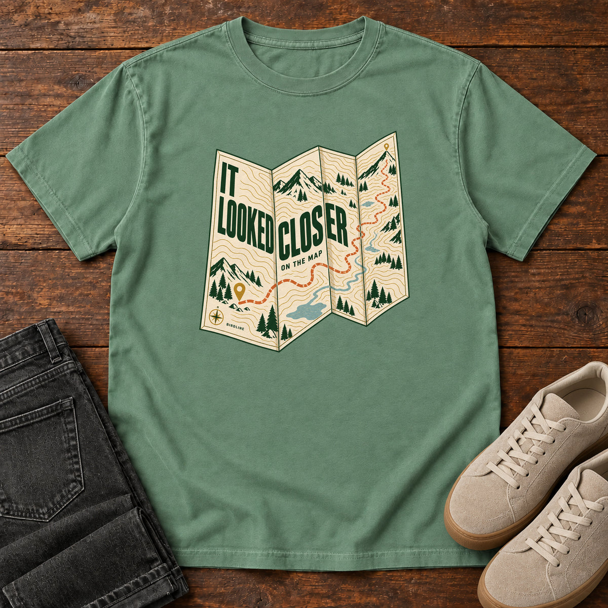 Light Green It Looked Closer on the Map T-Shirt with a folded cream map graphic, green lettering and an orange route, front flat lay on wood.