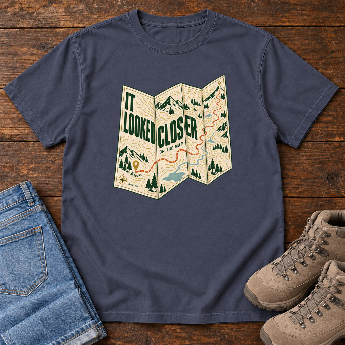 Navy It Looked Closer on the Map T-Shirt with a folded cream map graphic, green lettering and an orange route, front flat lay on wood.