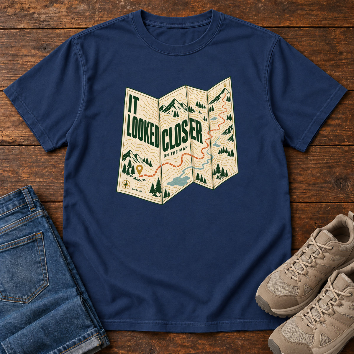 True Navy It Looked Closer on the Map T-Shirt with a folded cream map graphic, green lettering and an orange route, front flat lay on wood.