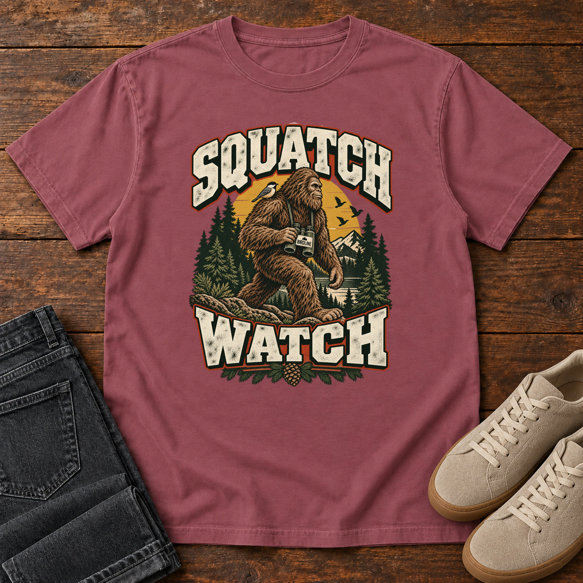 Squatch Watch T-Shirt in Brick, front laid flat on wood, featuring a walking Sasquatch holding binoculars among pine trees, framed by large cream lettering.