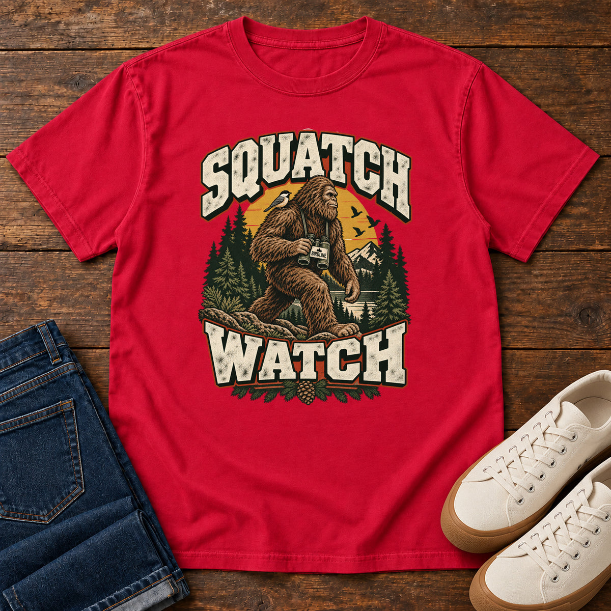 Squatch Watch T-Shirt in Red, front laid flat on wood, featuring a walking Sasquatch holding binoculars among pine trees, framed by large cream lettering.