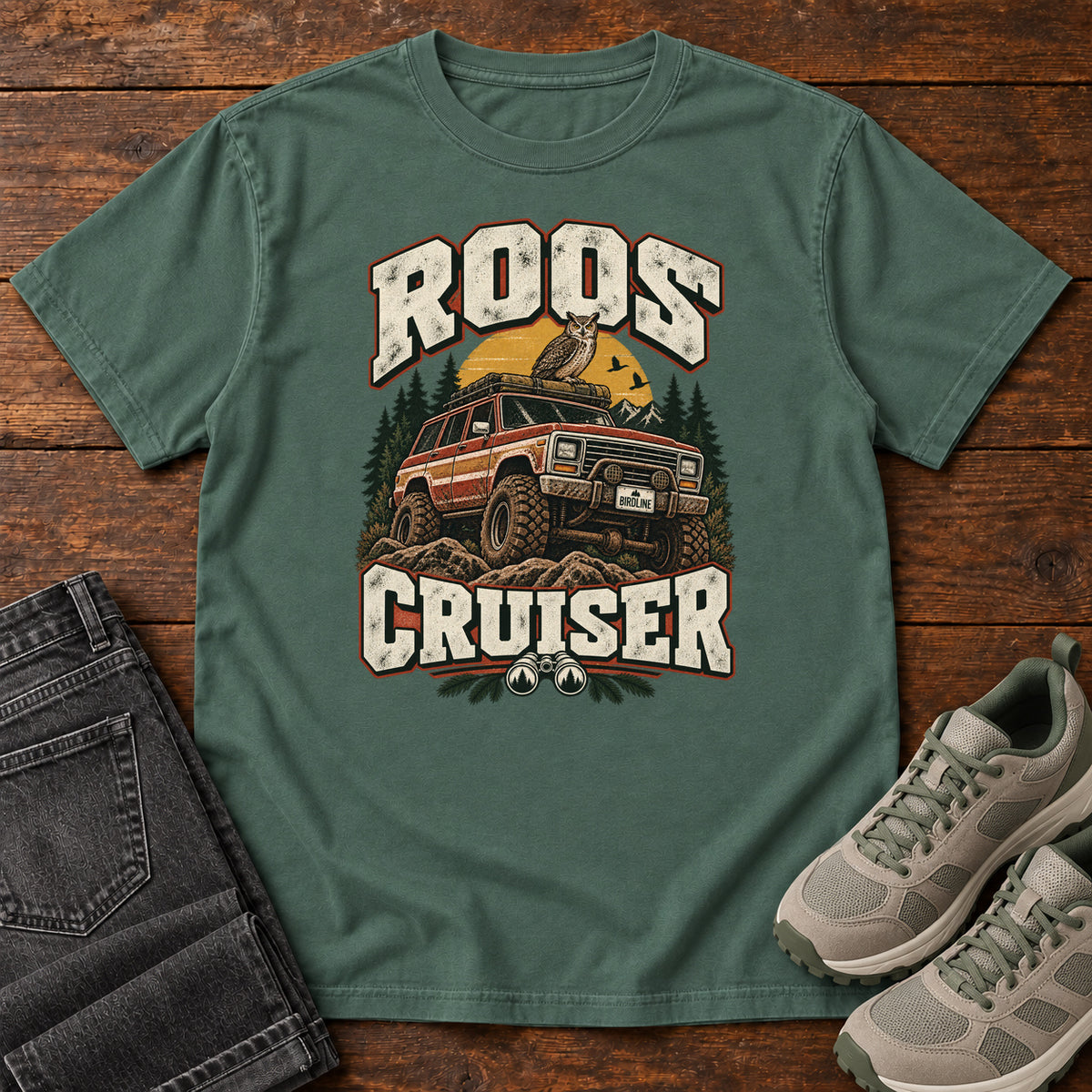 Blue Spruce Roos Cruiser T-Shirt with a bird perched on an off-road vehicle between distressed cream Roos Cruiser lettering, front flat lay on wood.