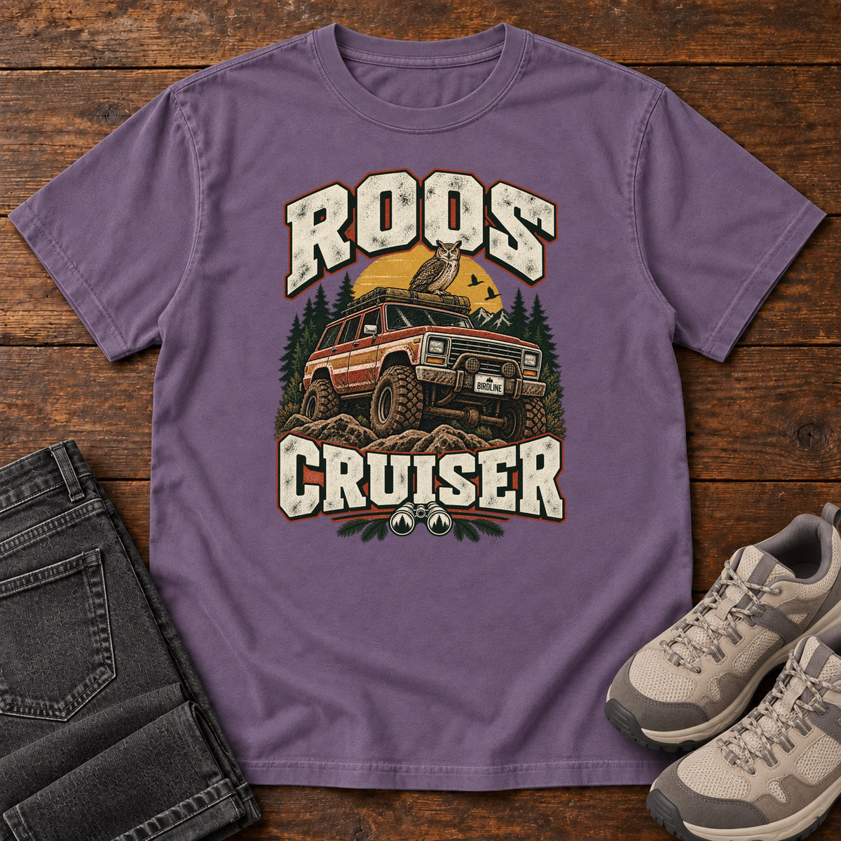 Grape Roos Cruiser T-Shirt with a bird perched on an off-road vehicle between distressed cream Roos Cruiser lettering, front flat lay on wood.