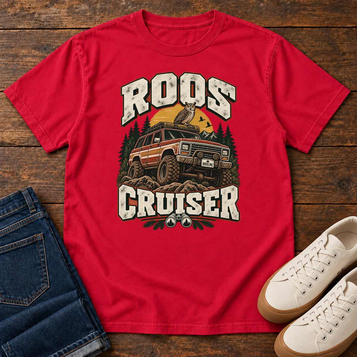 Red Roos Cruiser T-Shirt with a bird perched on an off-road vehicle between distressed cream Roos Cruiser lettering, front flat lay on wood.