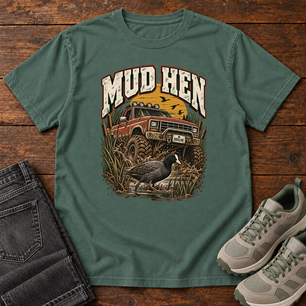 Blue Spruce Mud Hen T-Shirt with a red off-road truck splashing through mud behind a dark waterbird and reeds; front flat lay on wood.