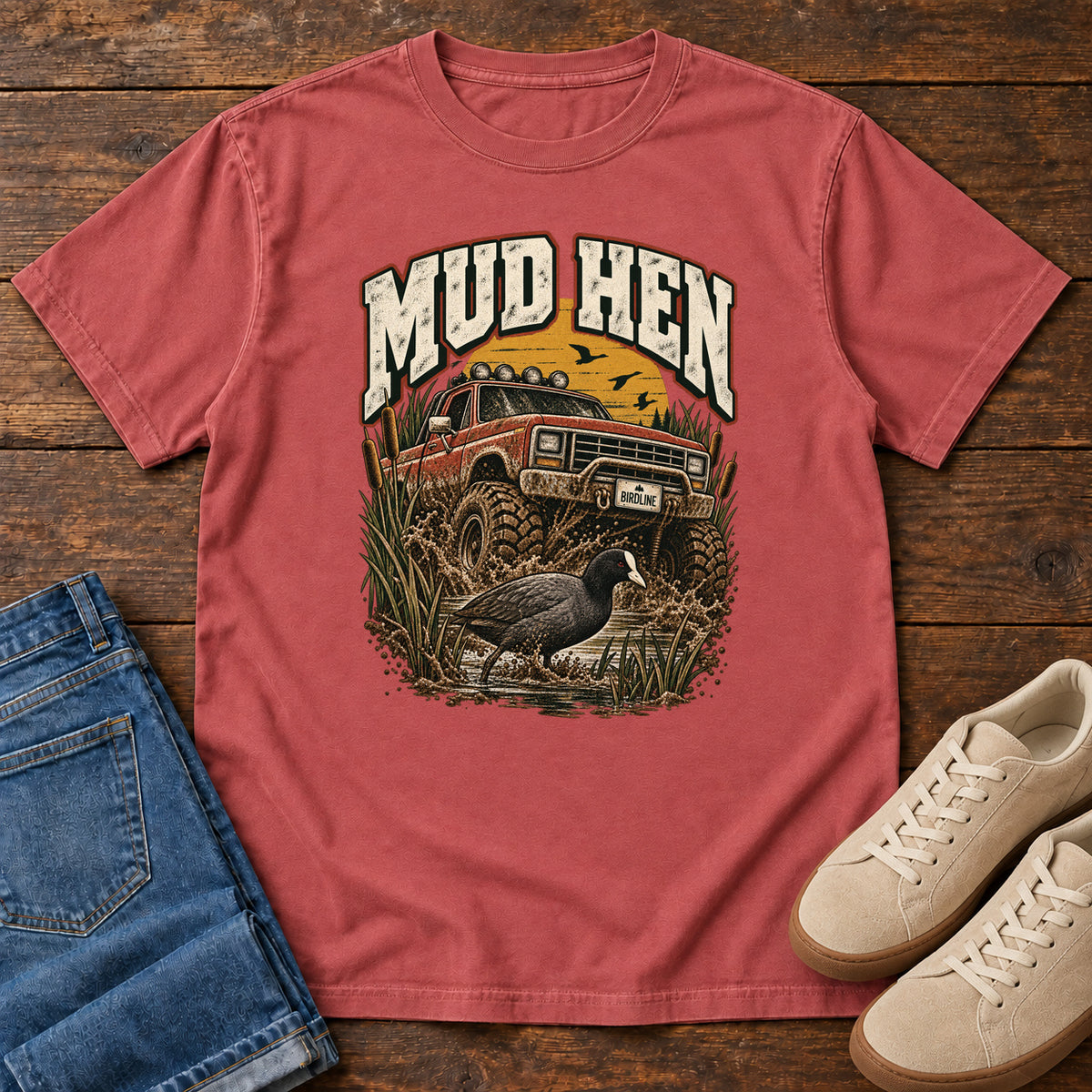 Crimson Mud Hen T-Shirt with a red off-road truck splashing through mud behind a dark waterbird and reeds; front flat lay on wood.