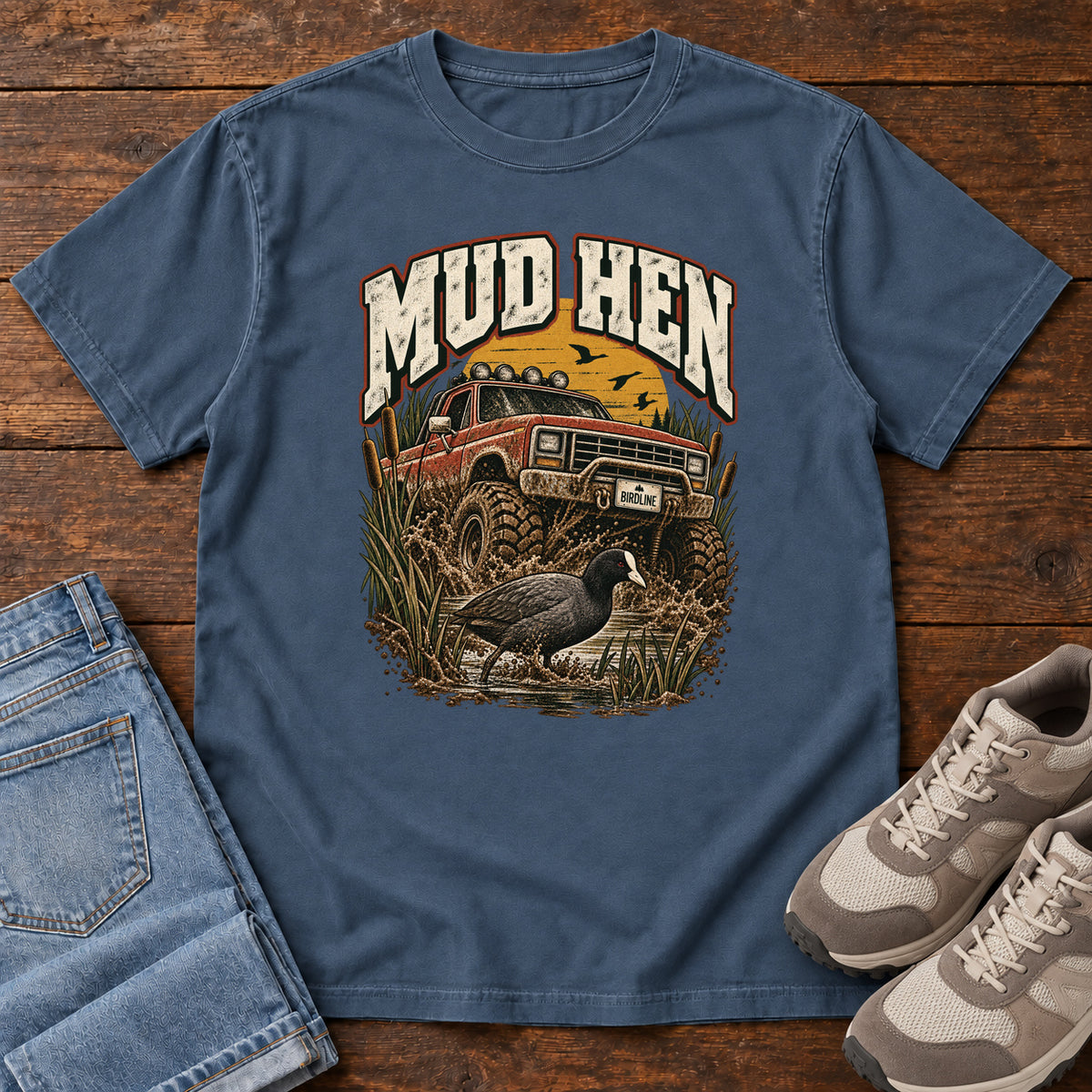 Midnight Mud Hen T-Shirt with a red off-road truck splashing through mud behind a dark waterbird and reeds; front flat lay on wood.