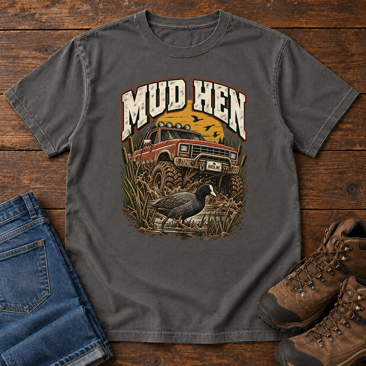 Pepper Mud Hen T-Shirt with a red off-road truck splashing through mud behind a dark waterbird and reeds; front flat lay on wood.
