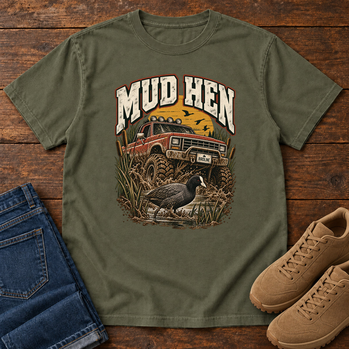 Sage Mud Hen T-Shirt with a red off-road truck splashing through mud behind a dark waterbird and reeds; front flat lay on wood.
