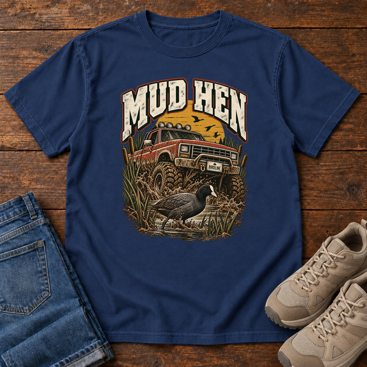 True Navy Mud Hen T-Shirt with a red off-road truck splashing through mud behind a dark waterbird and reeds; front flat lay on wood.
