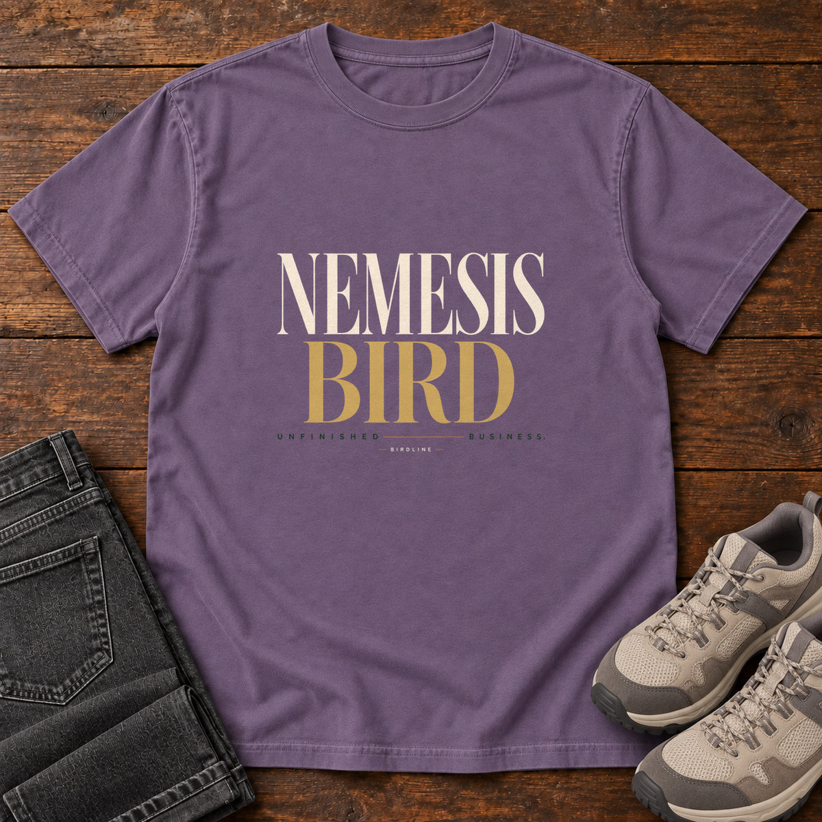 Grape Nemesis Bird T-Shirt with cream-and-gold serif lettering, front flat lay on wood.