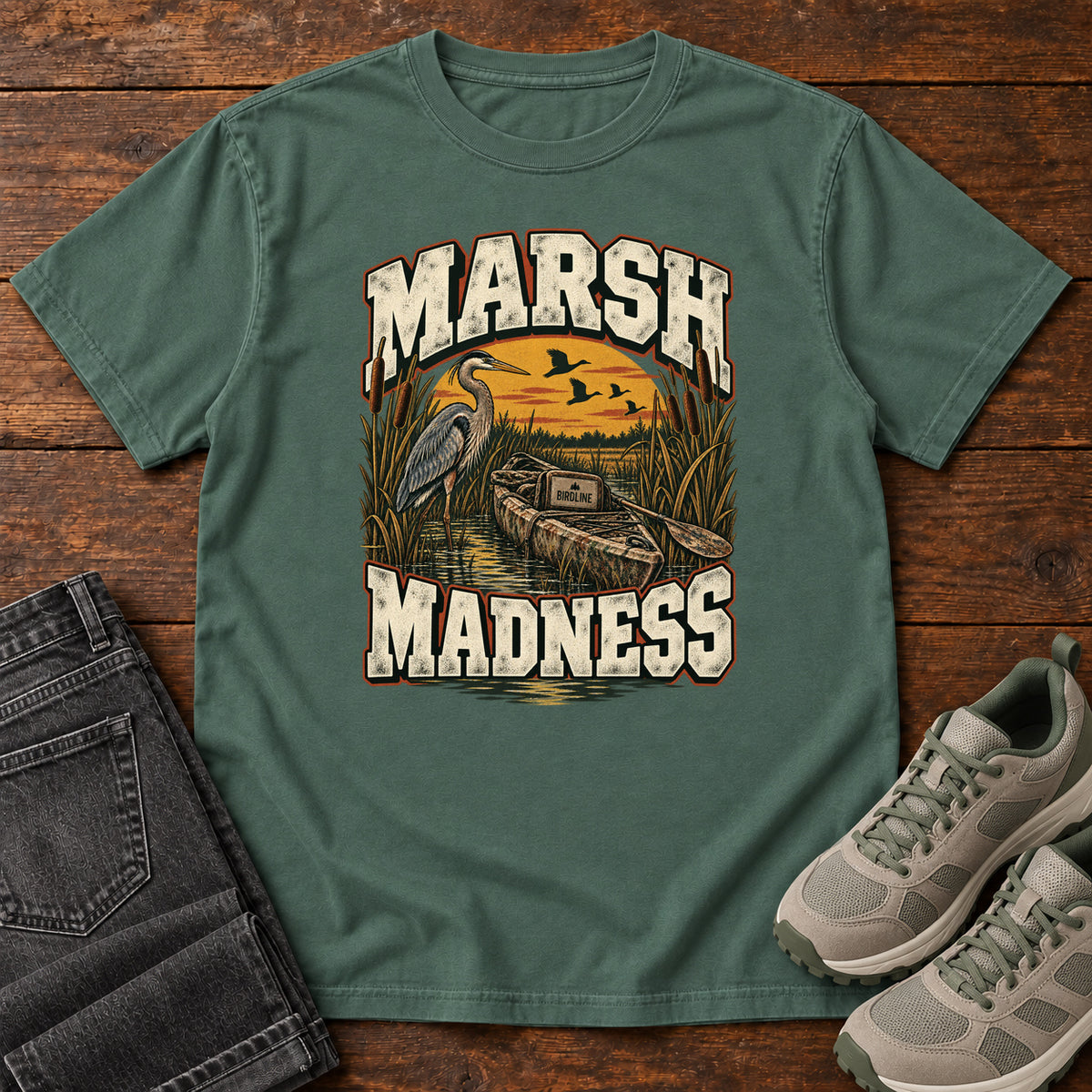 Blue Spruce Marsh Madness T-Shirt with a heron and canoe among marsh reeds beneath an orange sky and flying birds; front flat lay on wood.