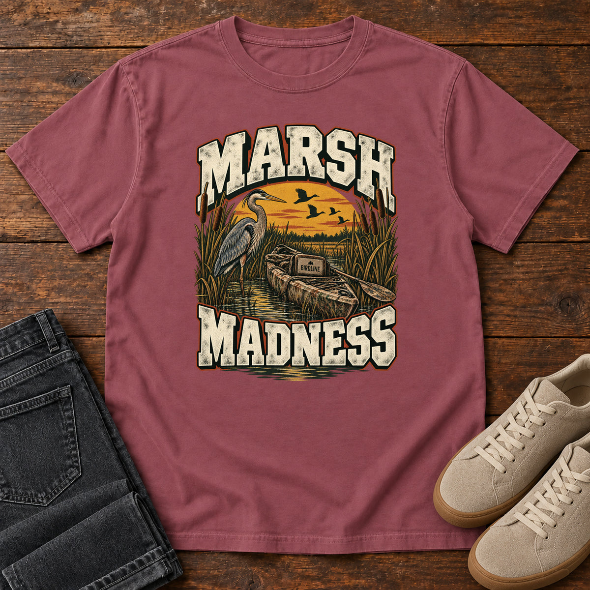 Brick Marsh Madness T-Shirt with a heron and canoe among marsh reeds beneath an orange sky and flying birds; front flat lay on wood.