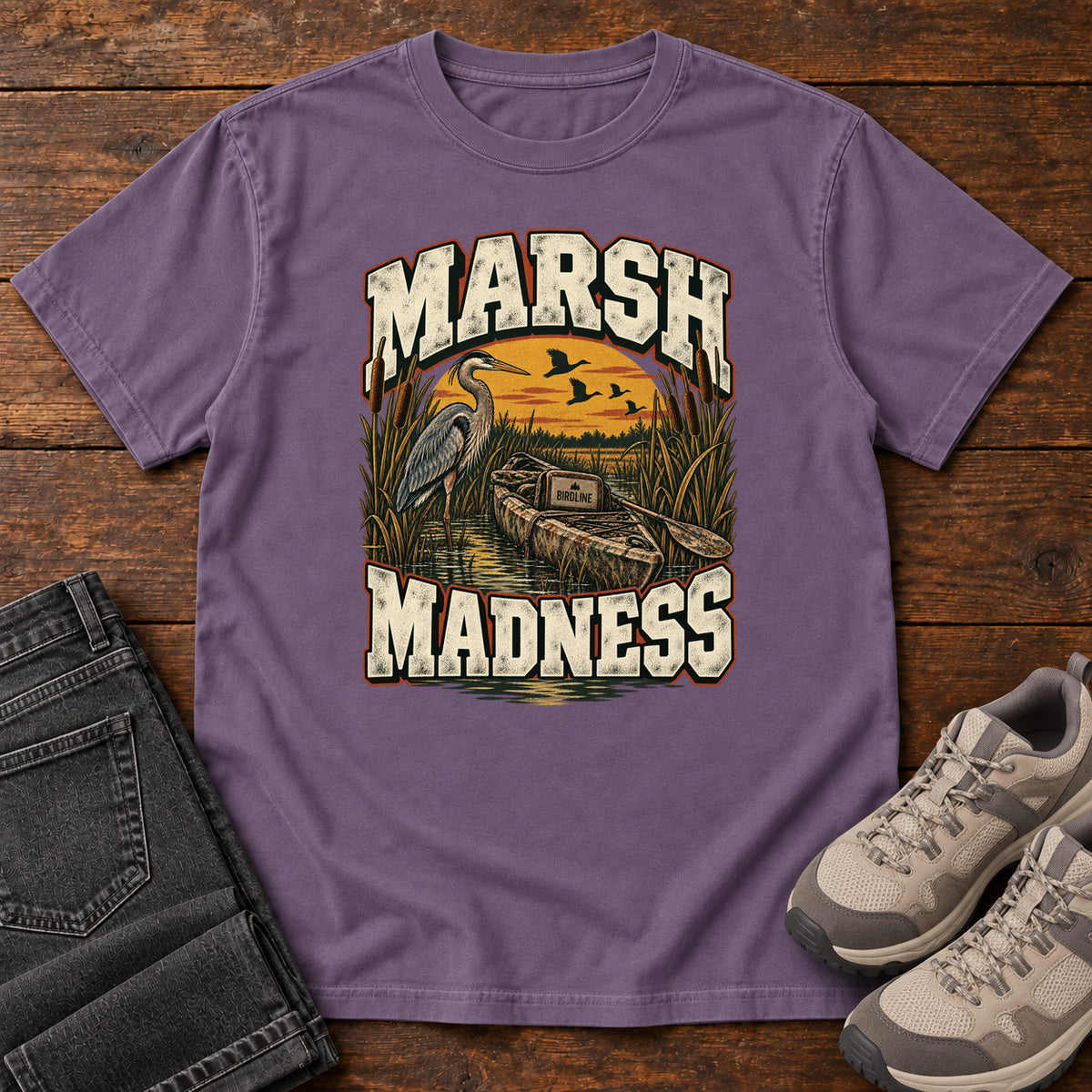 Grape Marsh Madness T-Shirt with a heron and canoe among marsh reeds beneath an orange sky and flying birds; front flat lay on wood.