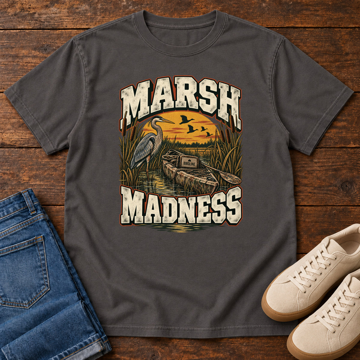 Graphite Marsh Madness T-Shirt with a heron and canoe among marsh reeds beneath an orange sky and flying birds; front flat lay on wood.