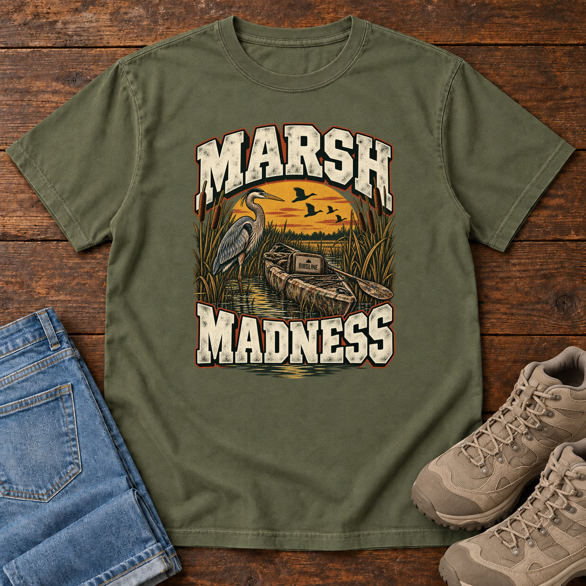 Hemp Marsh Madness T-Shirt with a heron and canoe among marsh reeds beneath an orange sky and flying birds; front flat lay on wood.