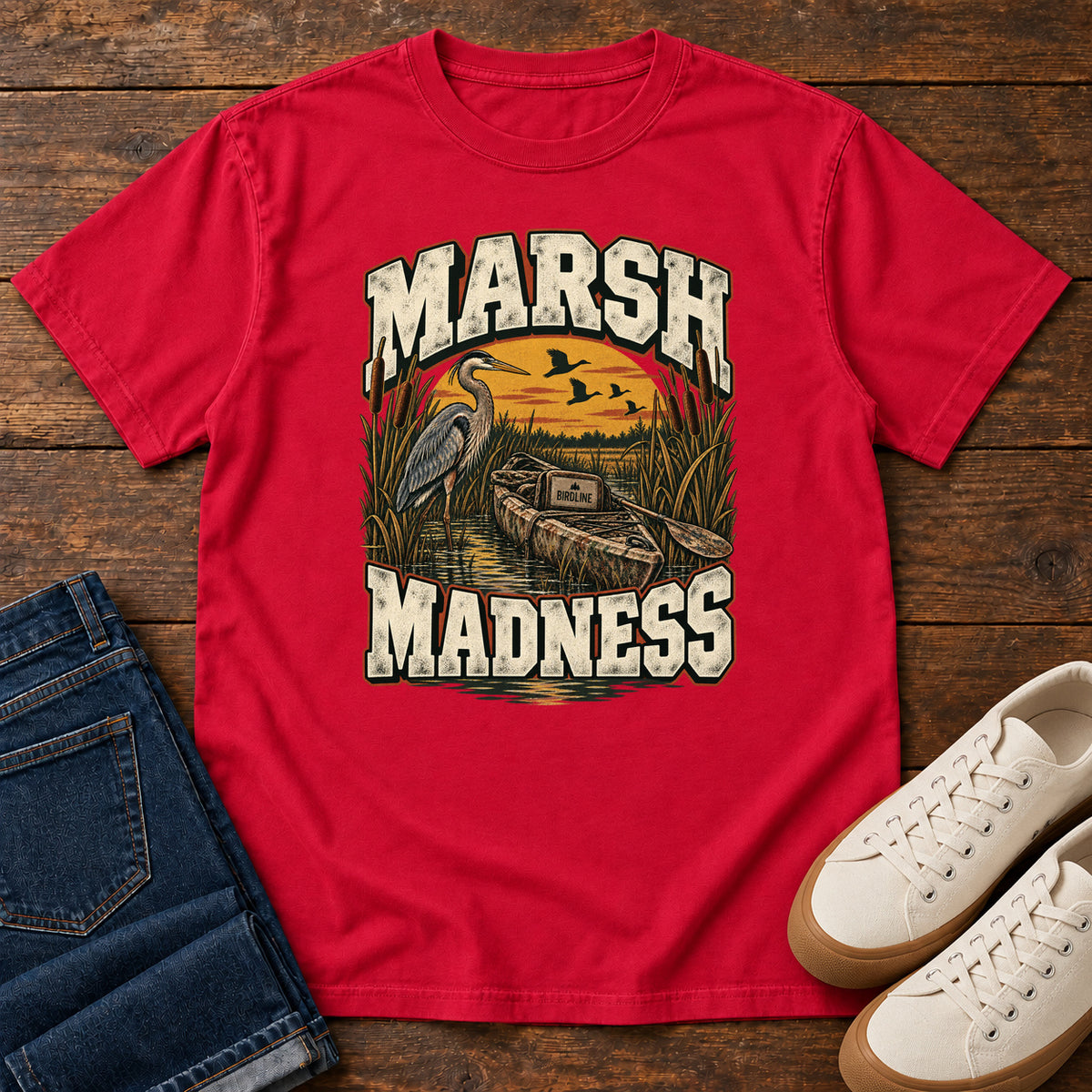 Red Marsh Madness T-Shirt with a heron and canoe among marsh reeds beneath an orange sky and flying birds; front flat lay on wood.