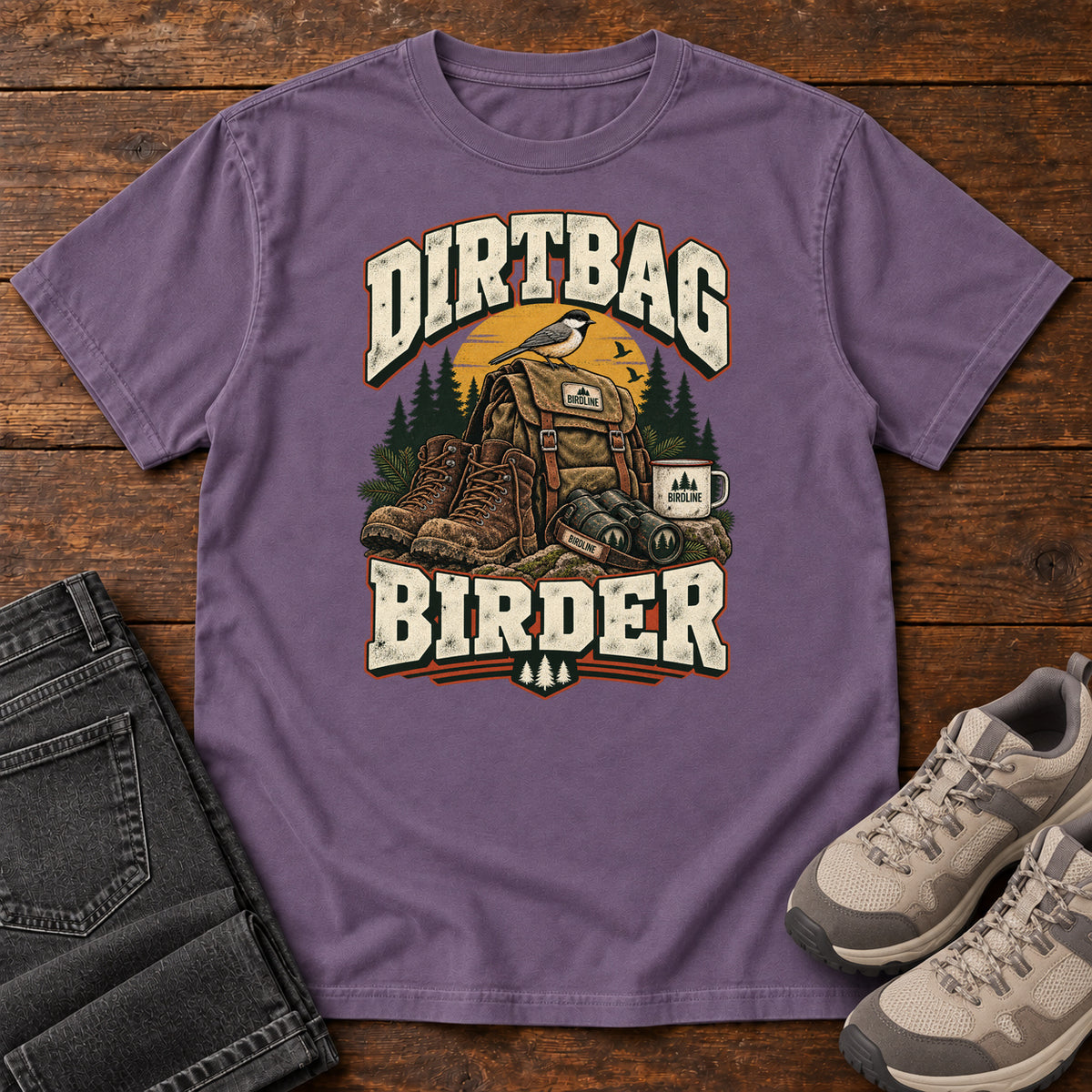 Grape Dirtbag Birder T-Shirt with hiking boots, a backpack, binoculars and a camping mug beneath cream lettering, front flat lay on wood.