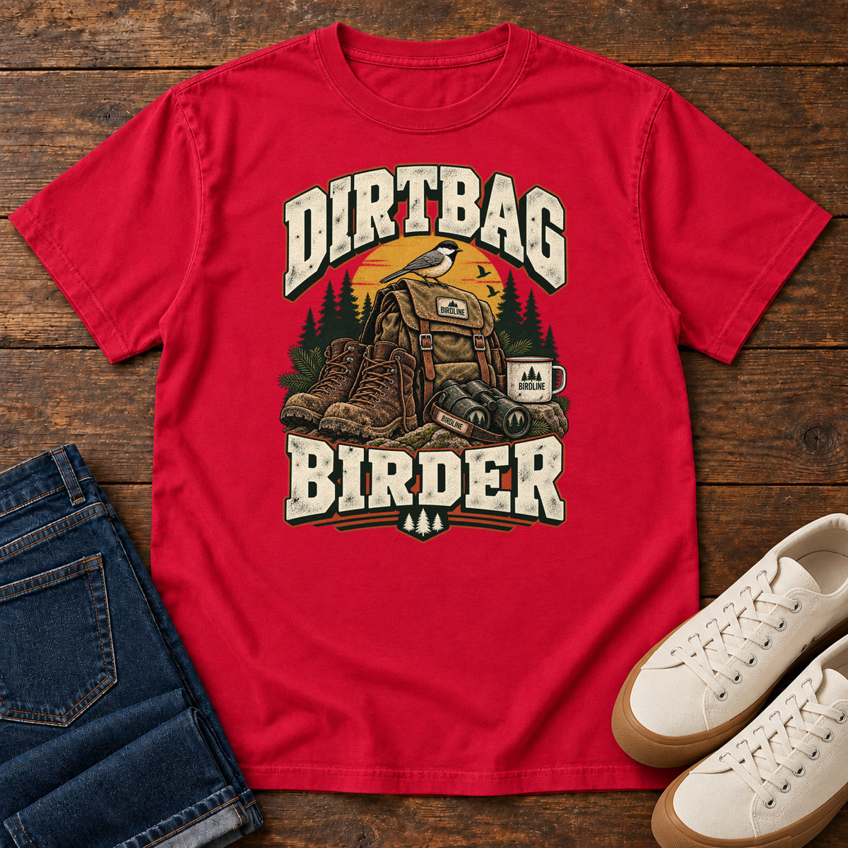 Red Dirtbag Birder T-Shirt with hiking boots, a backpack, binoculars and a camping mug beneath cream lettering, front flat lay on wood.