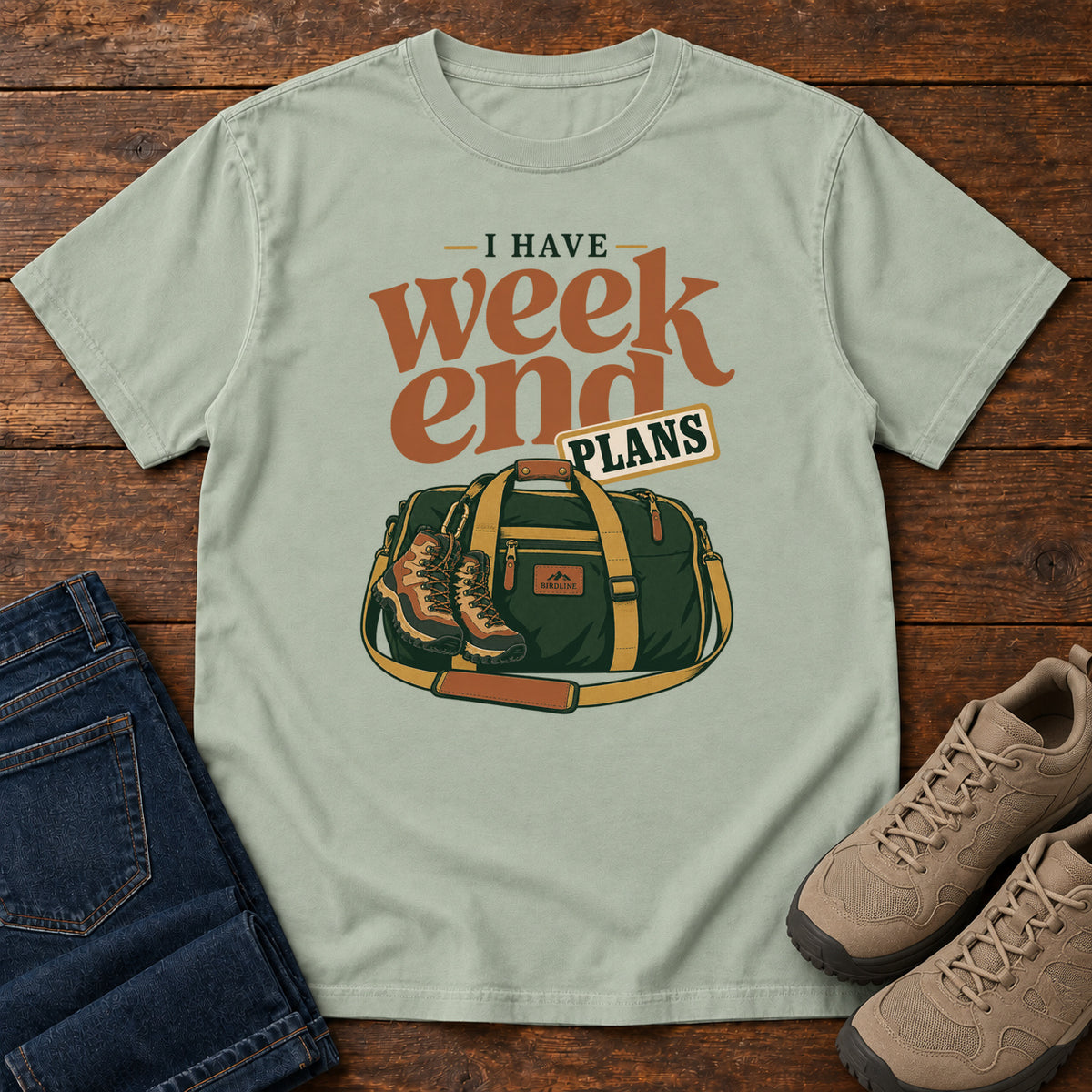 Bay I Have Weekend Plans T-Shirt with a green duffel bag and hiking boots beneath rust-orange Weekend lettering, front flat lay on wood.