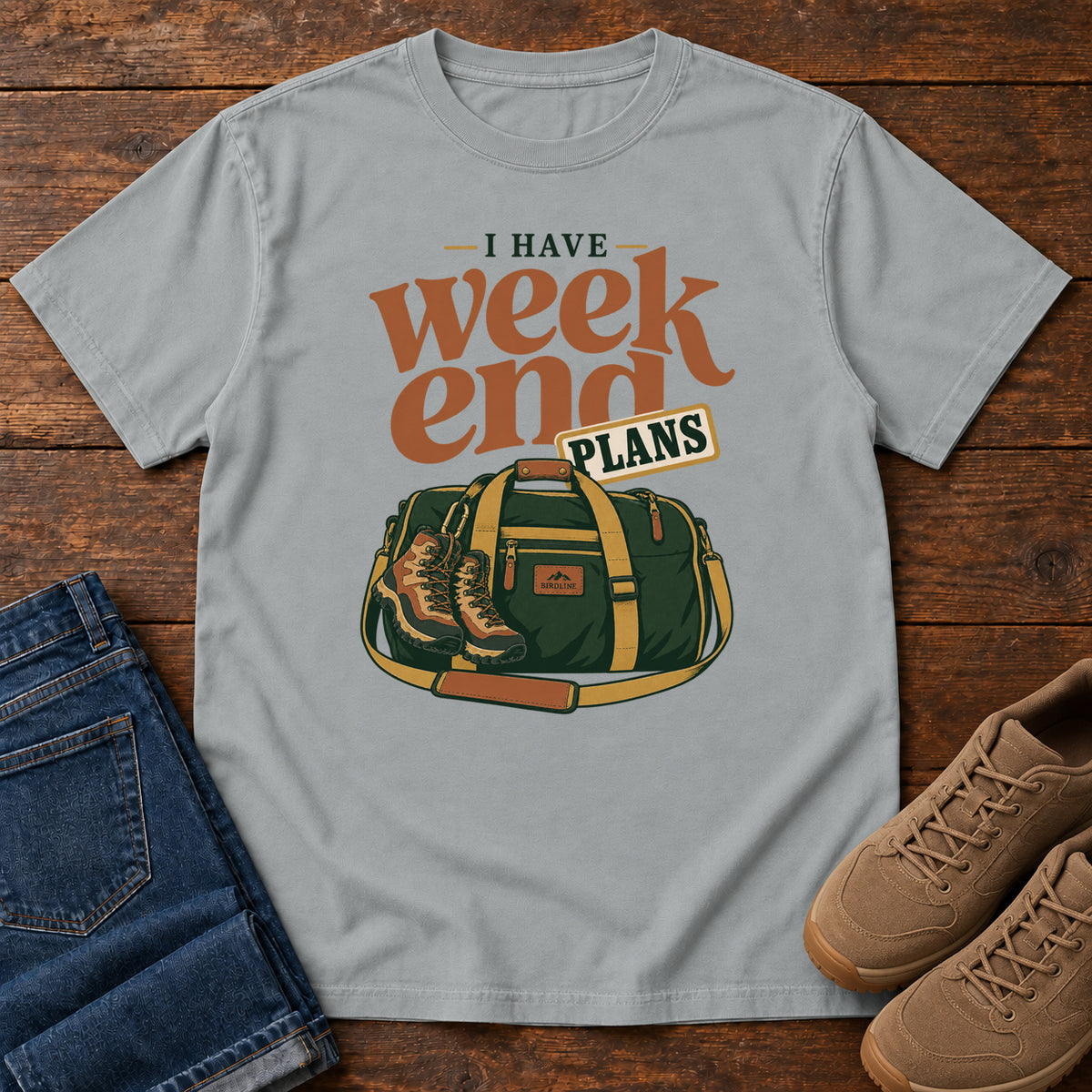 Granite I Have Weekend Plans T-Shirt with a green duffel bag and hiking boots beneath rust-orange Weekend lettering, front flat lay on wood.