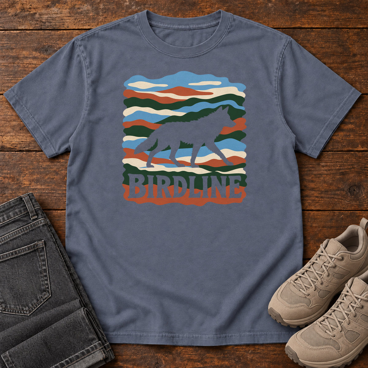 Denim Birdline Wolf Modern T-Shirt with a wolf silhouette over wavy multicolor stripes and Birdline lettering, front flat lay on wood.