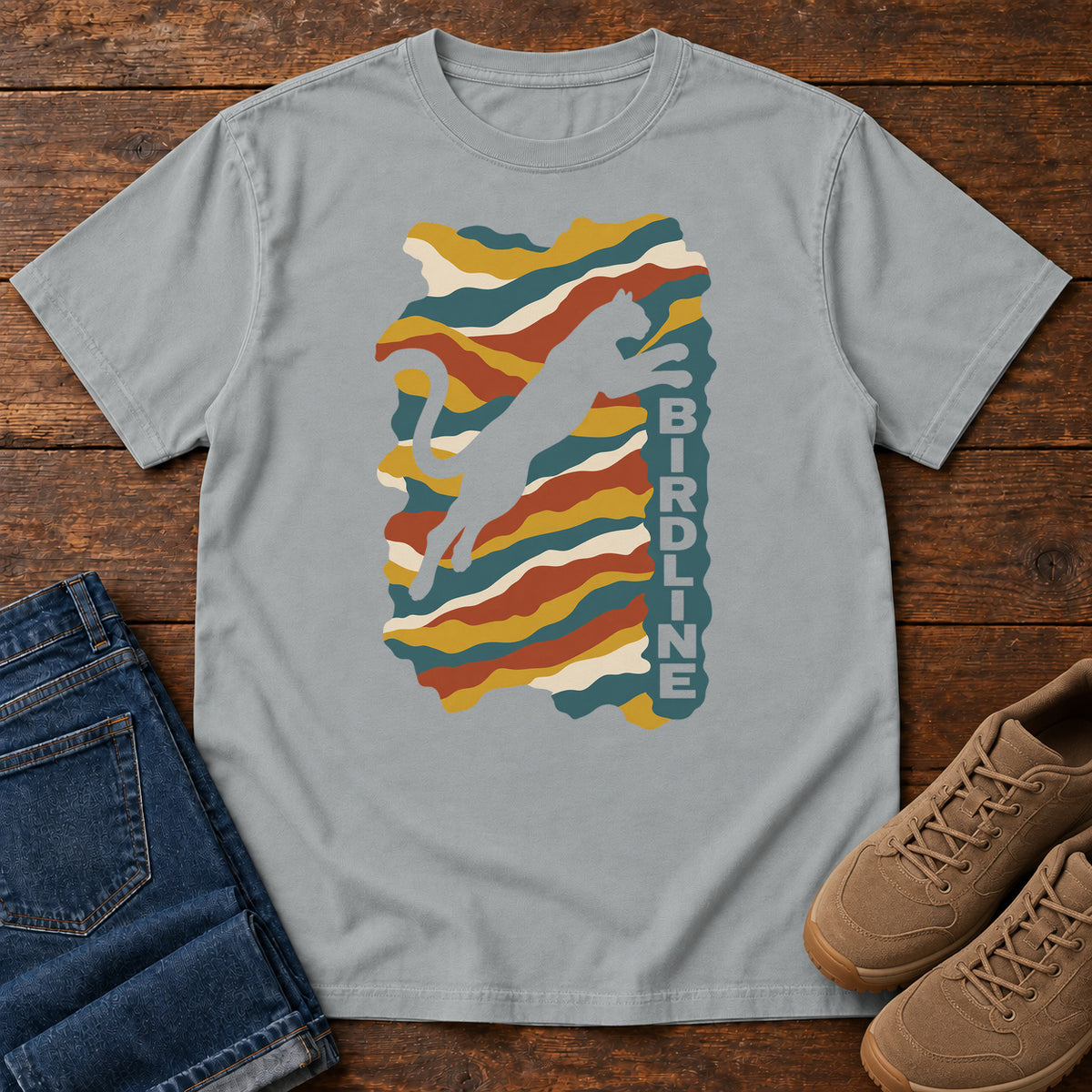 Granite Birdline Leaping Cat T-Shirt with a leaping cat silhouette over colorful wavy stripes and vertical Birdline lettering; front flat lay on wood.