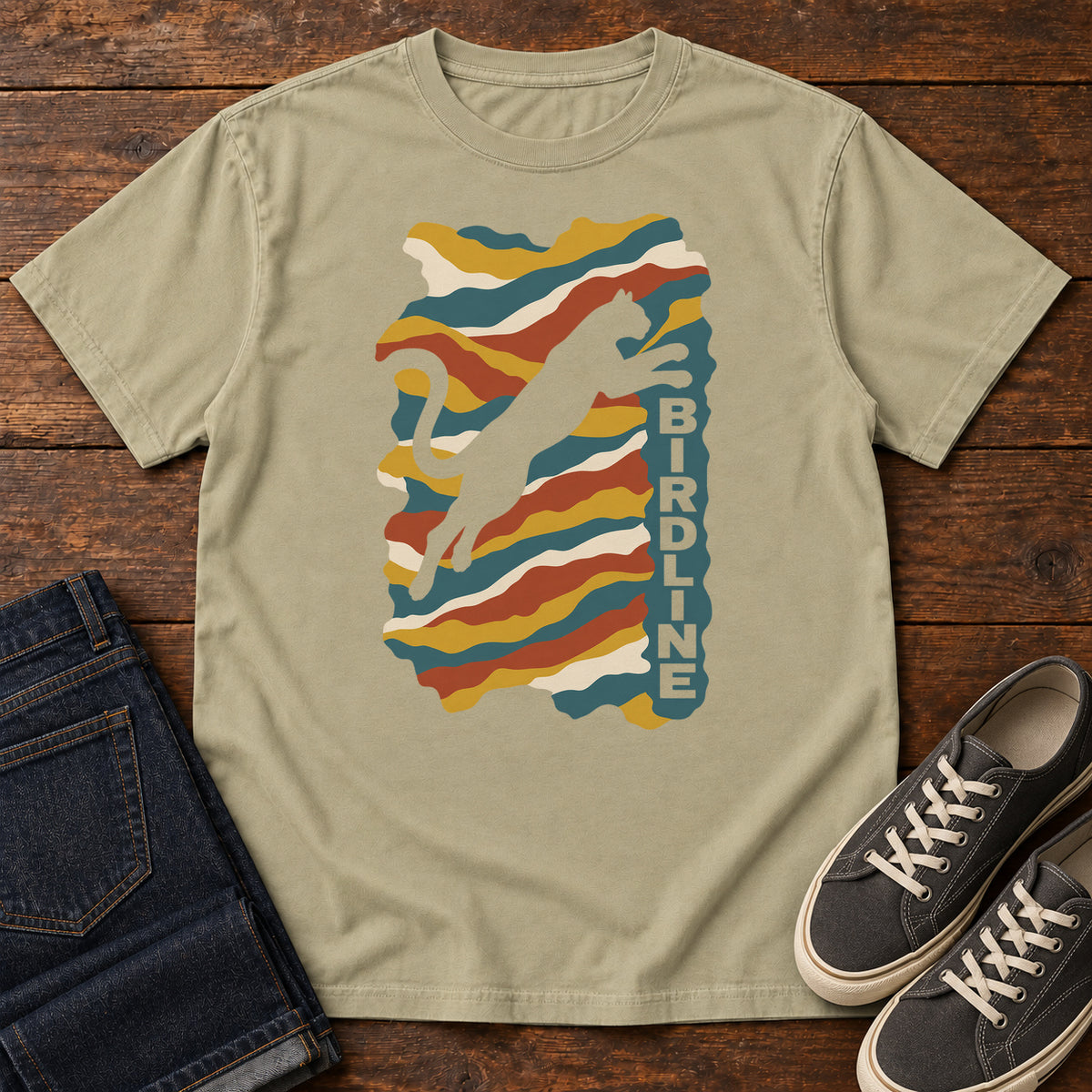 Khaki Birdline Leaping Cat T-Shirt with a leaping cat silhouette over colorful wavy stripes and vertical Birdline lettering; front flat lay on wood.