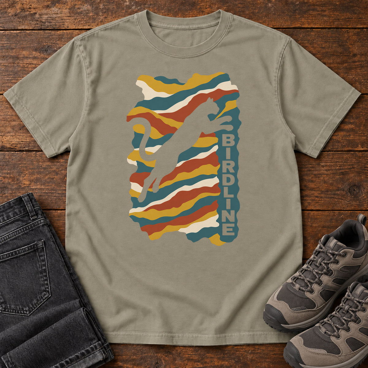 Sandstone Birdline Leaping Cat T-Shirt with a leaping cat silhouette over colorful wavy stripes and vertical Birdline lettering; front flat lay on wood.