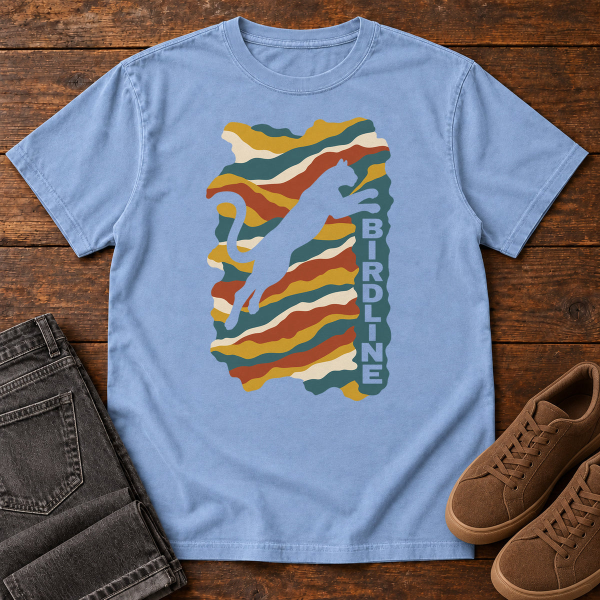 Washed Denim Birdline Leaping Cat T-Shirt with a leaping cat silhouette over colorful wavy stripes and vertical Birdline lettering; front flat lay on wood.