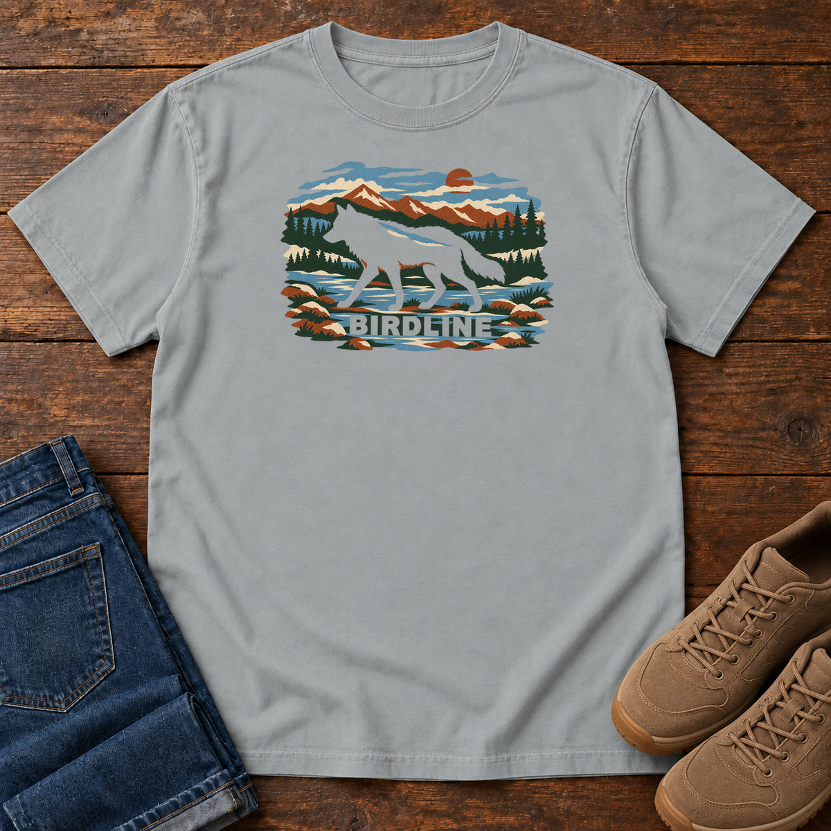 Granite Birdline Wolf Mountain T-Shirt with a wolf silhouette in a mountain, forest and river scene with Birdline lettering, front flat lay on wood.