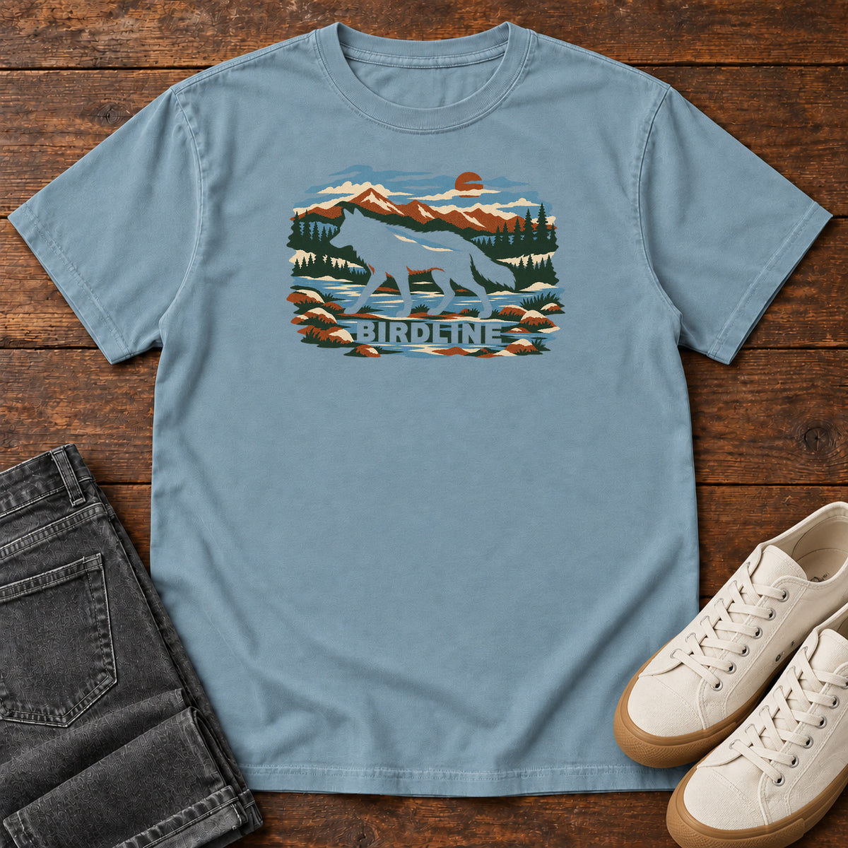 Ice Blue Birdline Wolf Mountain T-Shirt with a wolf silhouette in a mountain, forest and river scene with Birdline lettering, front flat lay on wood.