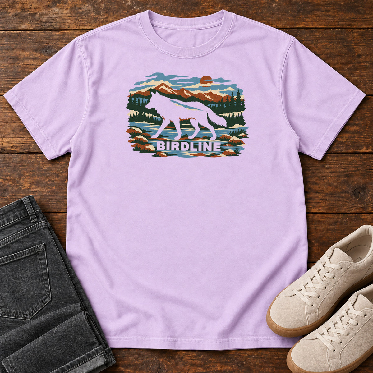 Orchid Birdline Wolf Mountain T-Shirt with a wolf silhouette in a mountain, forest and river scene with Birdline lettering, front flat lay on wood.