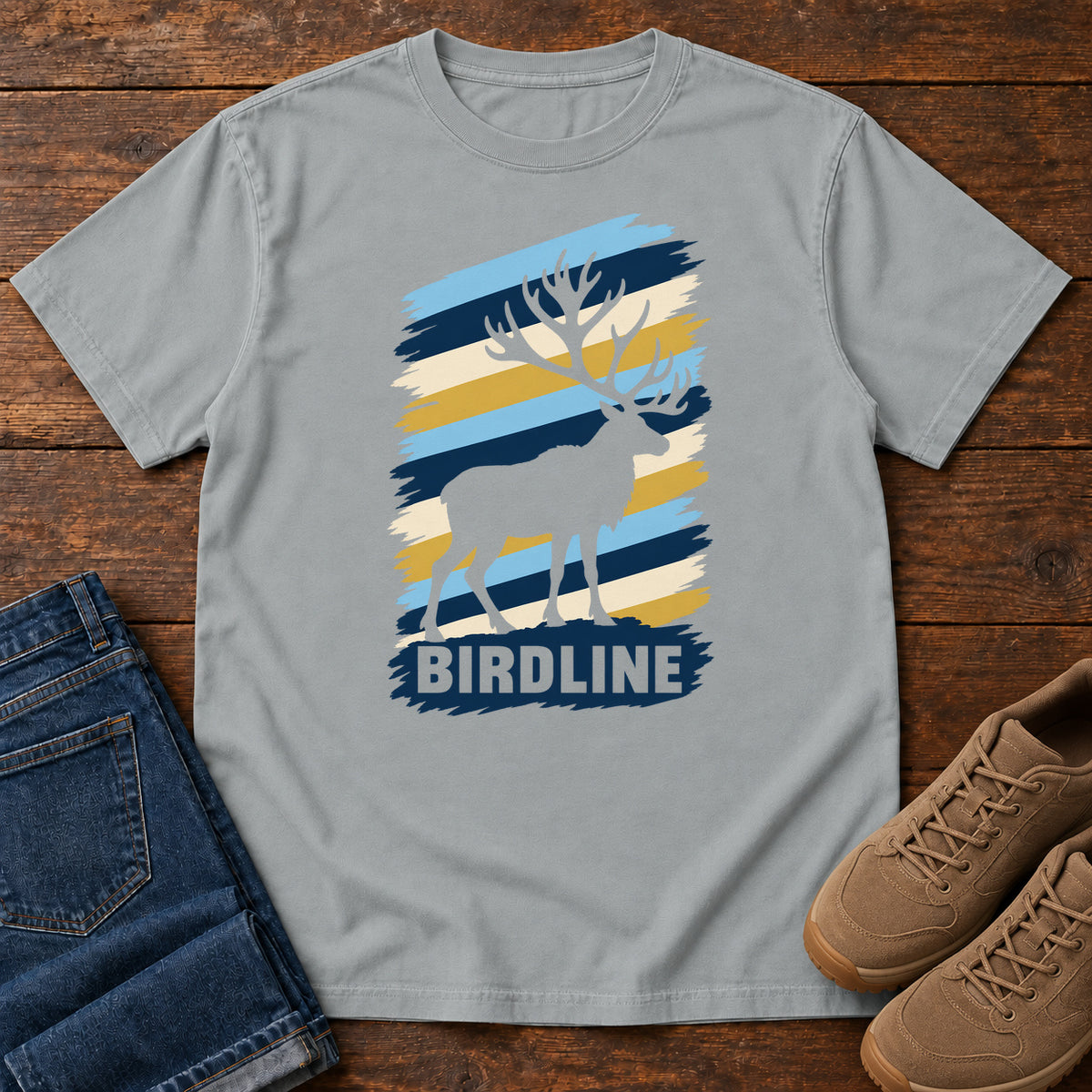 Birdline Elk Stripes T-Shirt in Granite, front laid flat on wood, featuring an elk silhouette against blue, cream and gold brushstroke stripes above BIRDLINE lettering.