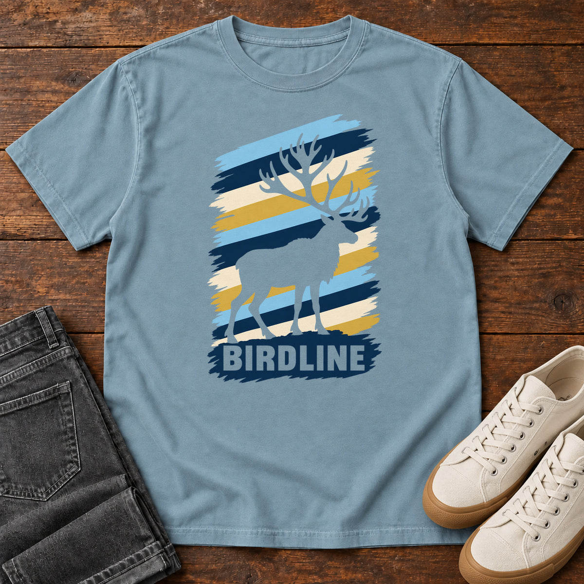 Birdline Elk Stripes T-Shirt in Ice Blue, front laid flat on wood, featuring an elk silhouette against blue, cream and gold brushstroke stripes above BIRDLINE lettering.
