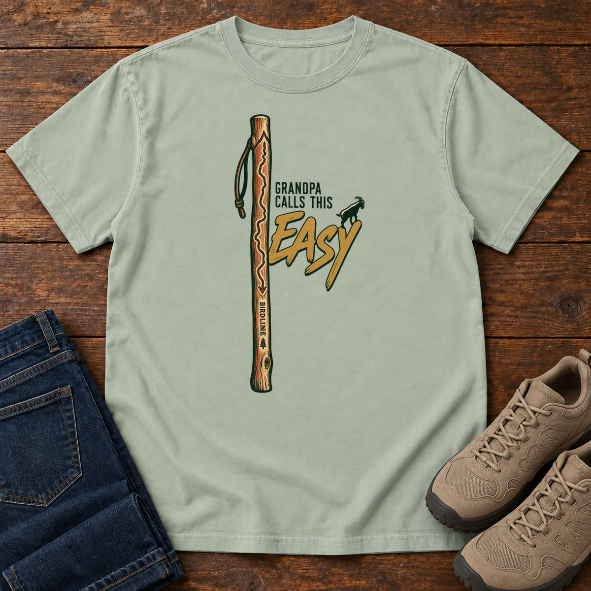 Bay Grandpa Calls This Easy T-Shirt with a wooden hiking stick beside green-and-gold lettering and a small goat silhouette; front flat lay on wood.