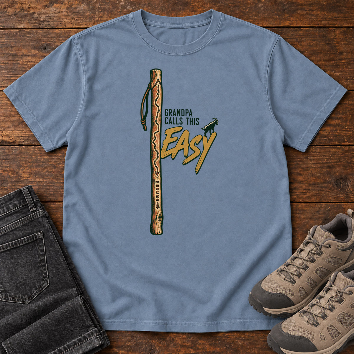 Blue Jean Grandpa Calls This Easy T-Shirt with a wooden hiking stick beside green-and-gold lettering and a small goat silhouette; front flat lay on wood.