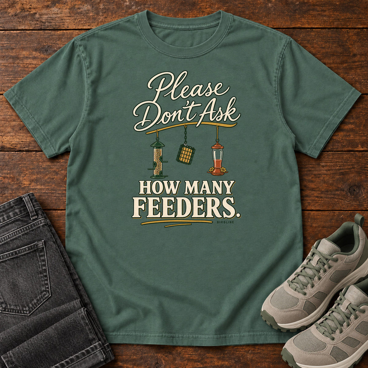 Blue Spruce Please Don't Ask How Many Feeders T-Shirt with cream lettering and three hanging bird feeders, front flat lay on wood.