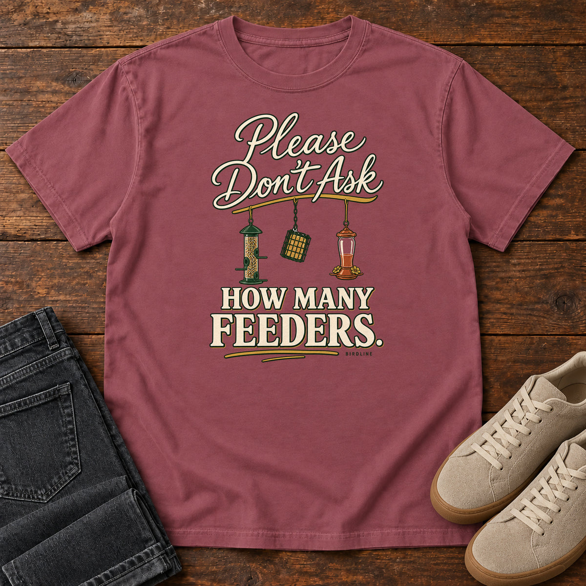 Brick Please Don't Ask How Many Feeders T-Shirt with cream lettering and three hanging bird feeders, front flat lay on wood.