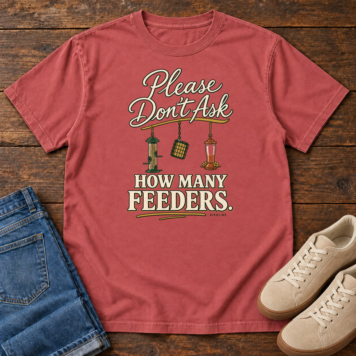 Crimson Please Don't Ask How Many Feeders T-Shirt with cream lettering and three hanging bird feeders, front flat lay on wood.