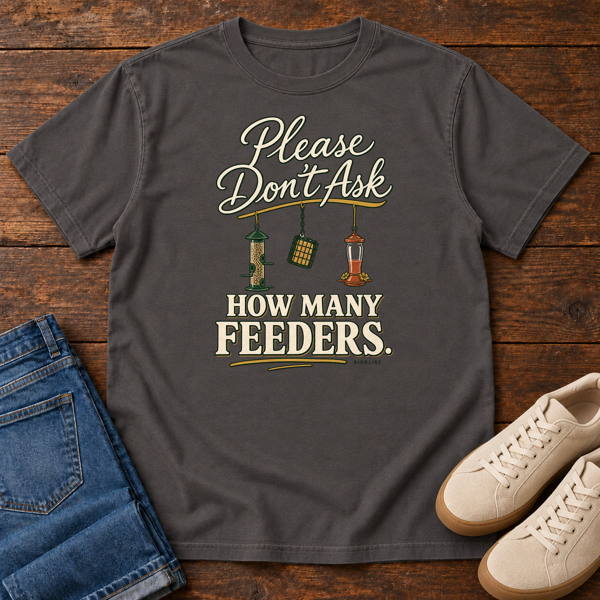 Graphite Please Don't Ask How Many Feeders T-Shirt with cream lettering and three hanging bird feeders, front flat lay on wood.