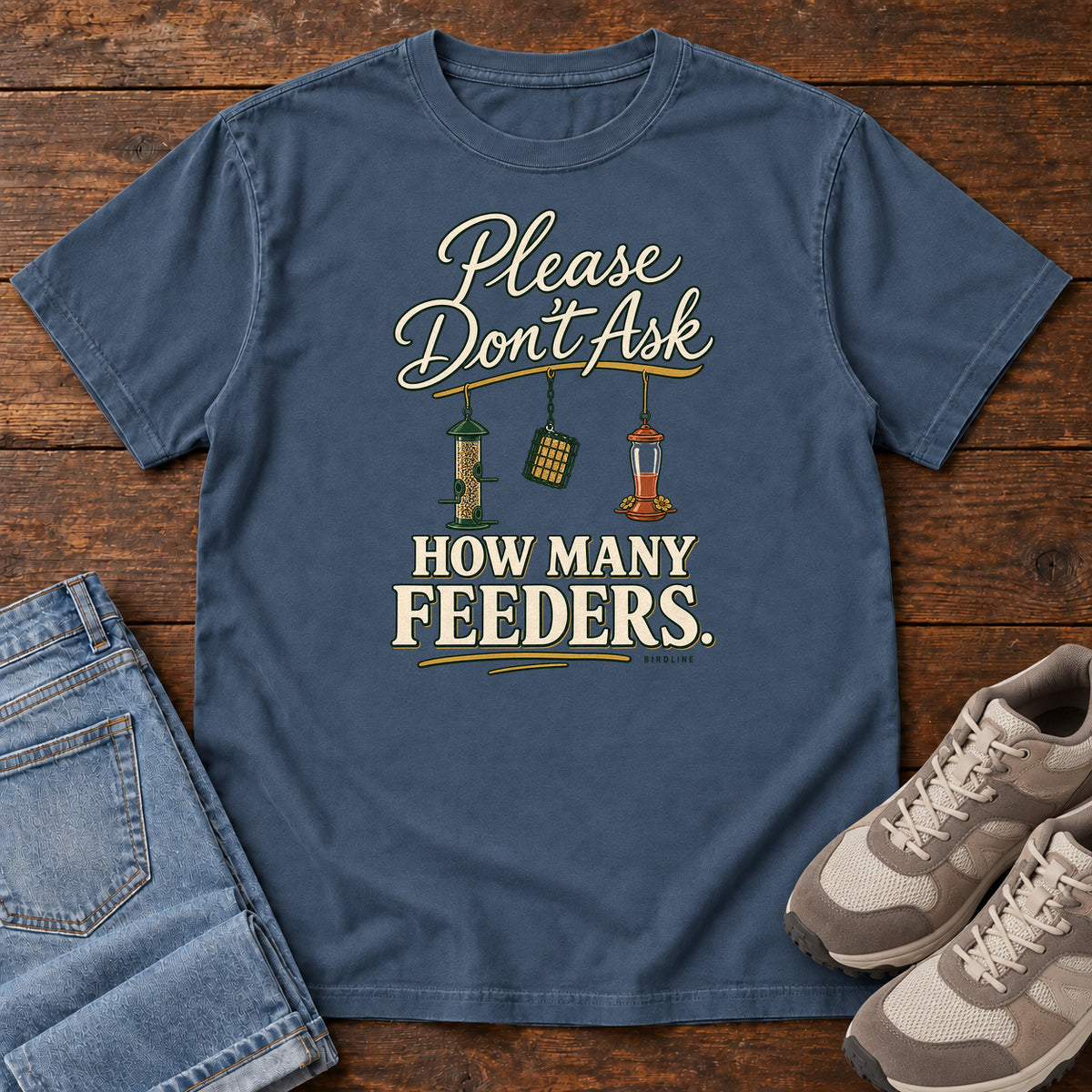 Midnight Please Don't Ask How Many Feeders T-Shirt with cream lettering and three hanging bird feeders, front flat lay on wood.