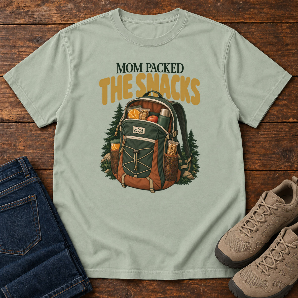Bay Mom Packed the Snacks T-Shirt with a snack-filled green backpack framed by evergreens and gold lettering; front flat lay on wood.