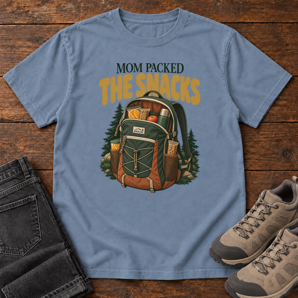 Blue Jean Mom Packed the Snacks T-Shirt with a snack-filled green backpack framed by evergreens and gold lettering; front flat lay on wood.