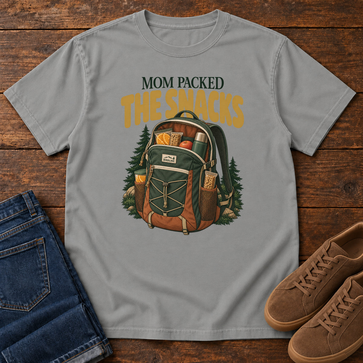 Grey Mom Packed the Snacks T-Shirt with a snack-filled green backpack framed by evergreens and gold lettering; front flat lay on wood.