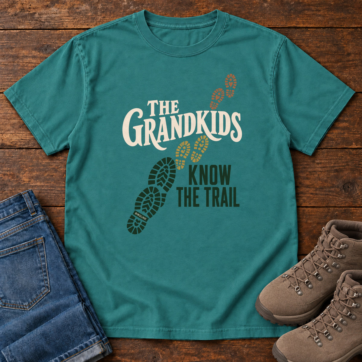 Emerald The Grandkids Know the Trail T-Shirt with cream and green lettering beside a trail of green, gold and brown boot prints, front flat lay on wood.