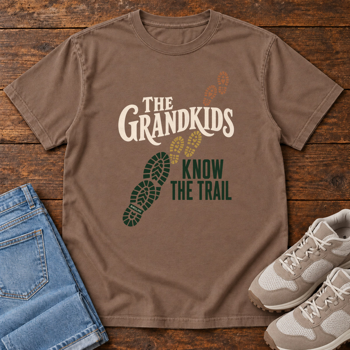 Espresso The Grandkids Know the Trail T-Shirt with cream and green lettering beside a trail of green, gold and brown boot prints, front flat lay on wood.