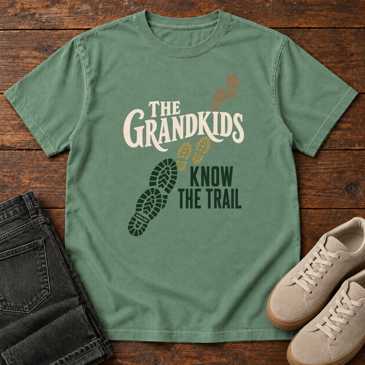 Light Green The Grandkids Know the Trail T-Shirt with cream and green lettering beside a trail of green, gold and brown boot prints, front flat lay on wood.